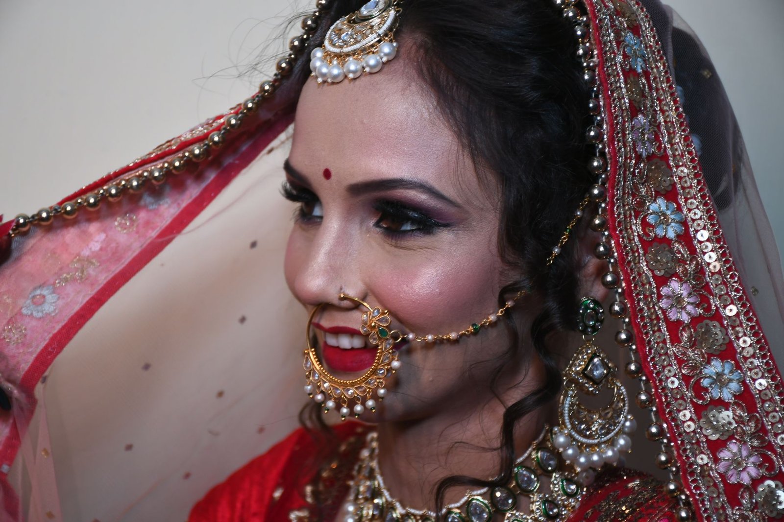 A beautiful Indian bride adorned with traditional jewelry and red lehenga, smiling and lifting her veil.