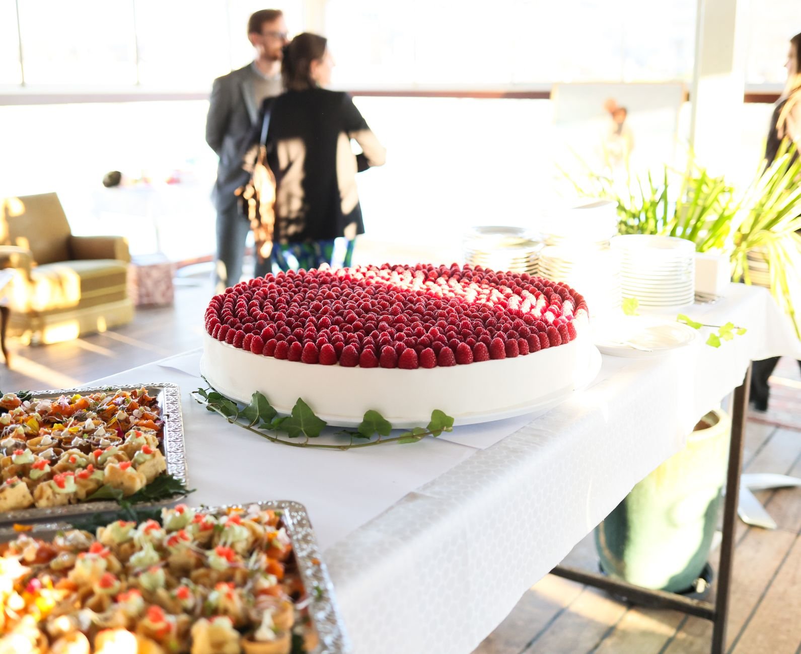 An elegant event table showcasing a large raspberry-topped cake and trays of hors d'oeuvres.