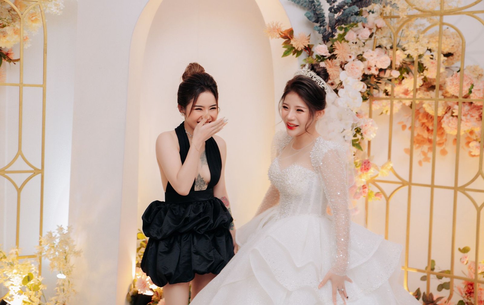Two women share a joyful moment in elegant dresses at a beautiful wedding setting adorned with flowers.