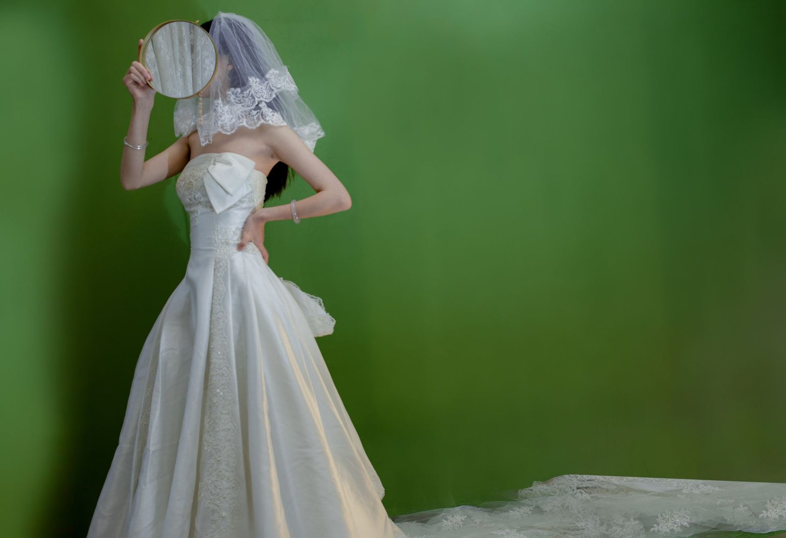Pengantin wanita elegan dalam gaun pengantin tanpa tali dan cadar berpose dengan cermin di setting studio.