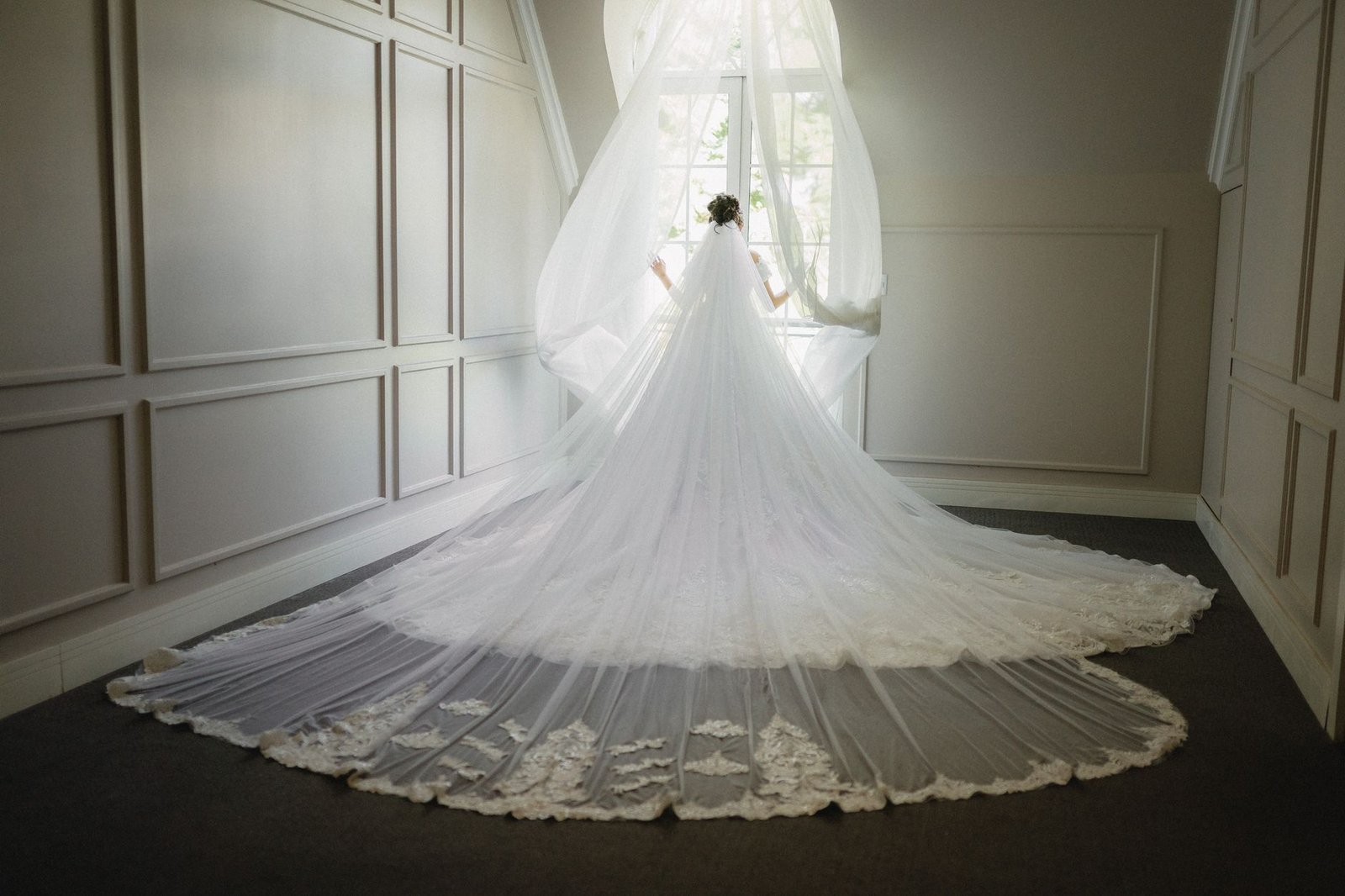 Seorang pengantin wanita yang menakjubkan dalam gaun elegan berdiri di dekat jendela yang terang benderang, menciptakan suasana yang tenang dan etereal.