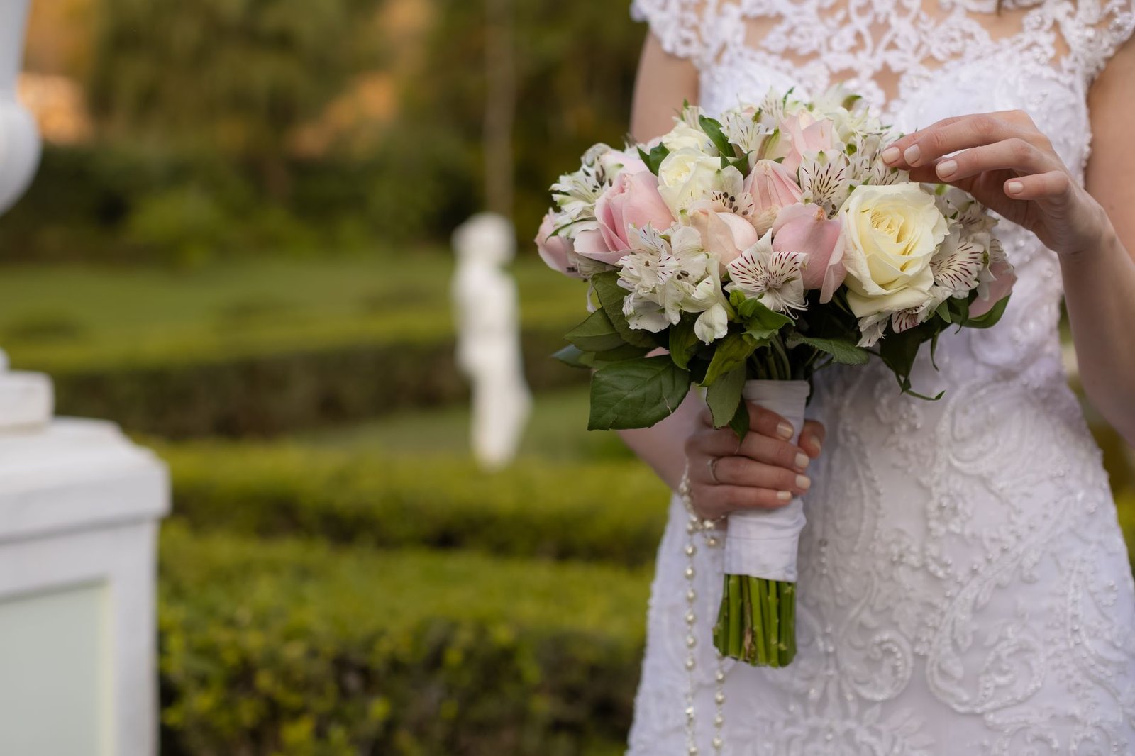 Seorang pengantin memegang buket bunga elegan dalam balutan gaun renda di luar ruangan dalam suasana taman.