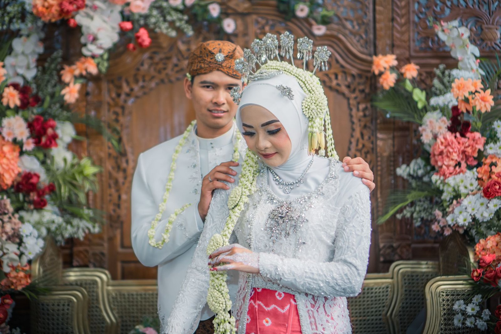 Potret cantik sepasang kekasih dalam balutan busana pengantin adat Indonesia, dikelilingi hiasan bunga semarak.