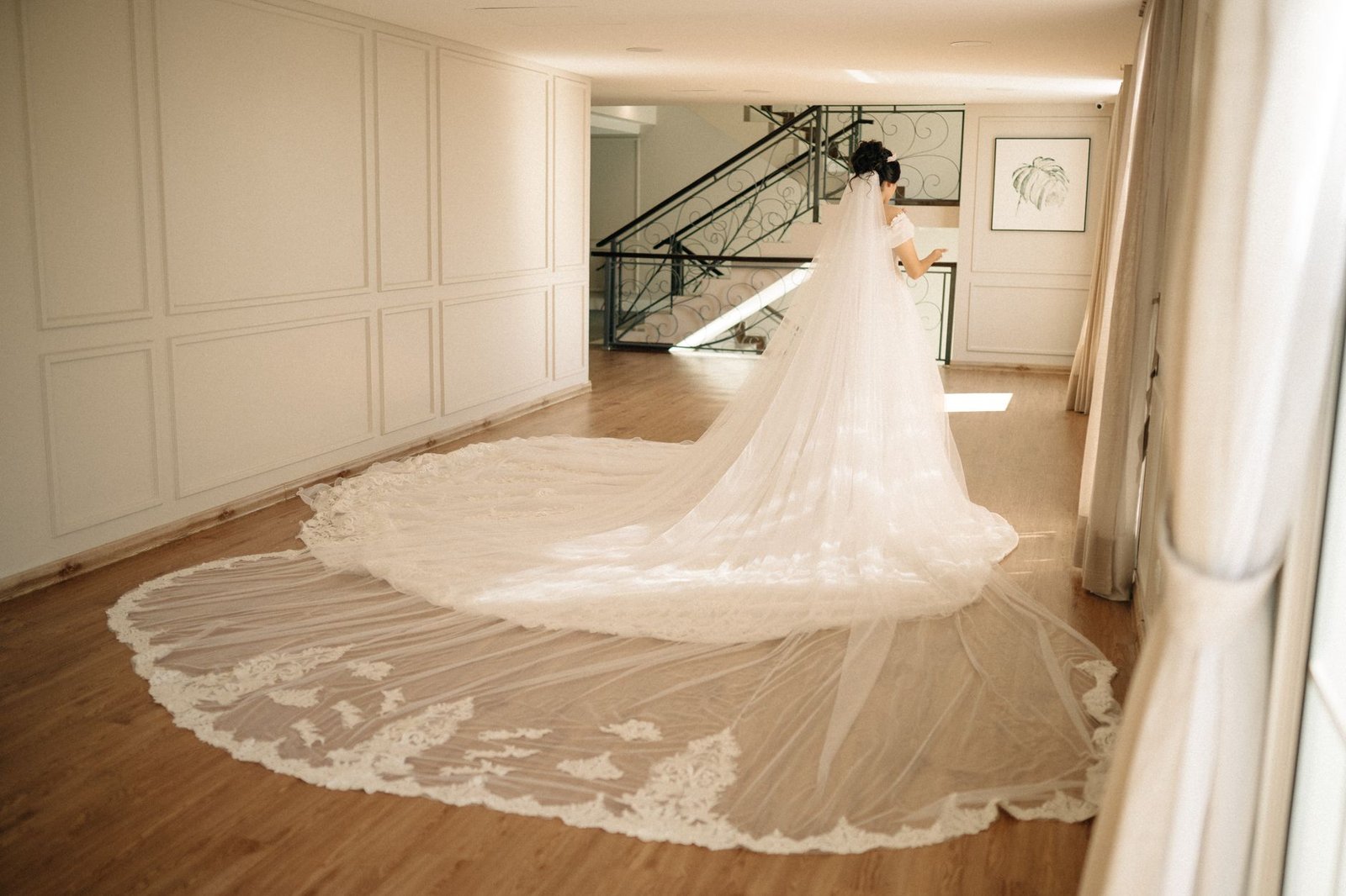 Seorang pengantin wanita dalam gaun putih dengan ekor panjang berdiri dengan elegan di dalam ruangan, memamerkan gaya pernikahan klasik.