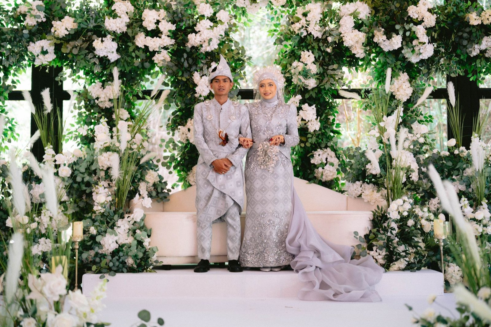 Sepasang pengantin dengan pakaian tradisional berpose di tengah rangkaian bunga yang rimbun pada upacara pernikahan mereka.