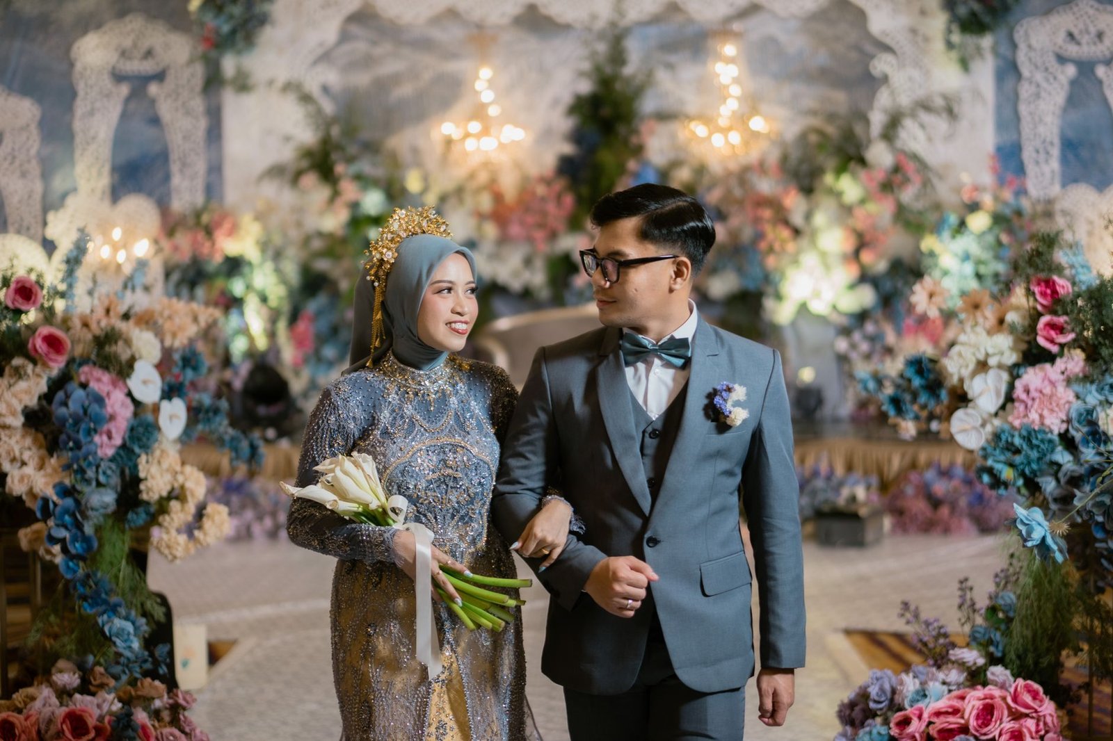 Pasangan pengantin yang menakjubkan dengan pakaian mewah dan latar belakang bunga yang semarak, merayakan pernikahan mereka di Indonesia.