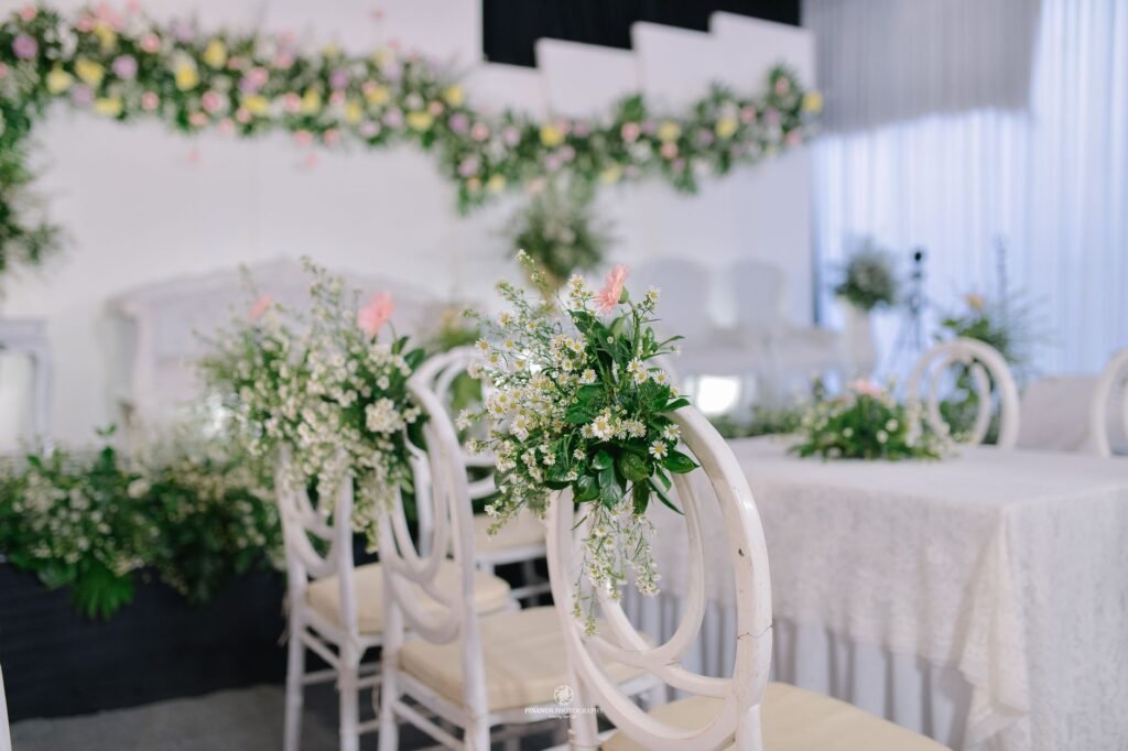 Tempat pernikahan elegan yang dilengkapi kursi putih dan rangkaian bunga yang rimbun, sempurna untuk acara yang tak terlupakan.