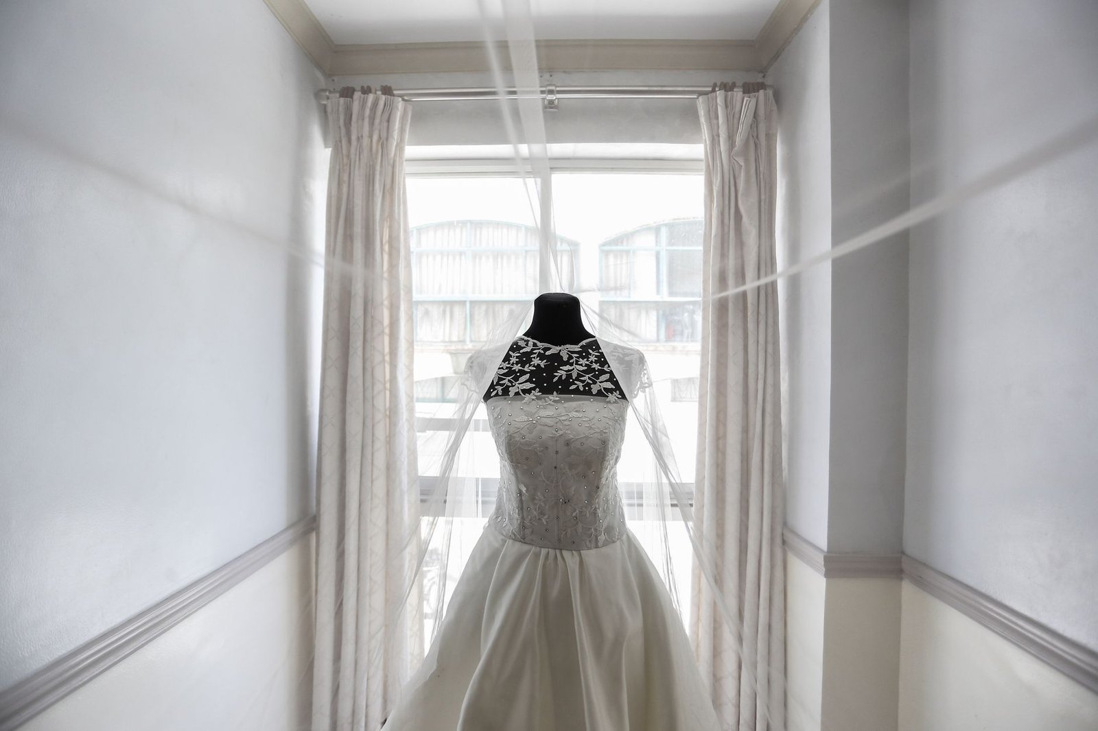 Gaun pengantin rajutan yang dipamerkan dengan anggun di atas manekin dekat jendela besar, menciptakan suasana yang lembut dan mewah.