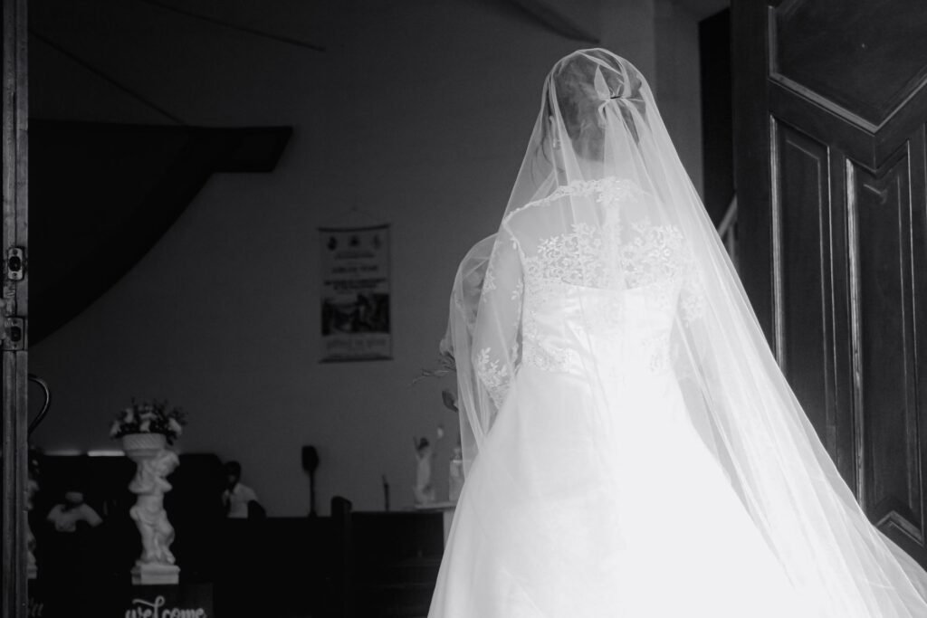 Pengantin wanita elegan dalam gaun putih dan slinga memasuki tempat pernikahan, diabadikan dalam suasana hitam dan putih.