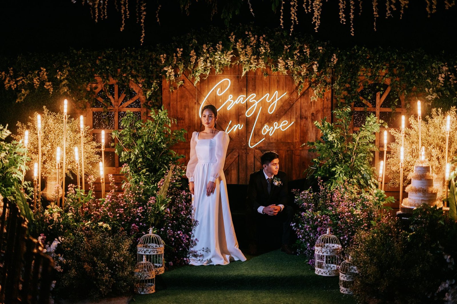 Sebuah adegan malam pernikahan yang didekorasi indah dengan pasangan pengantin baru mengenakan busana yang elegan dan dikelilingi vegetasi yang subur.