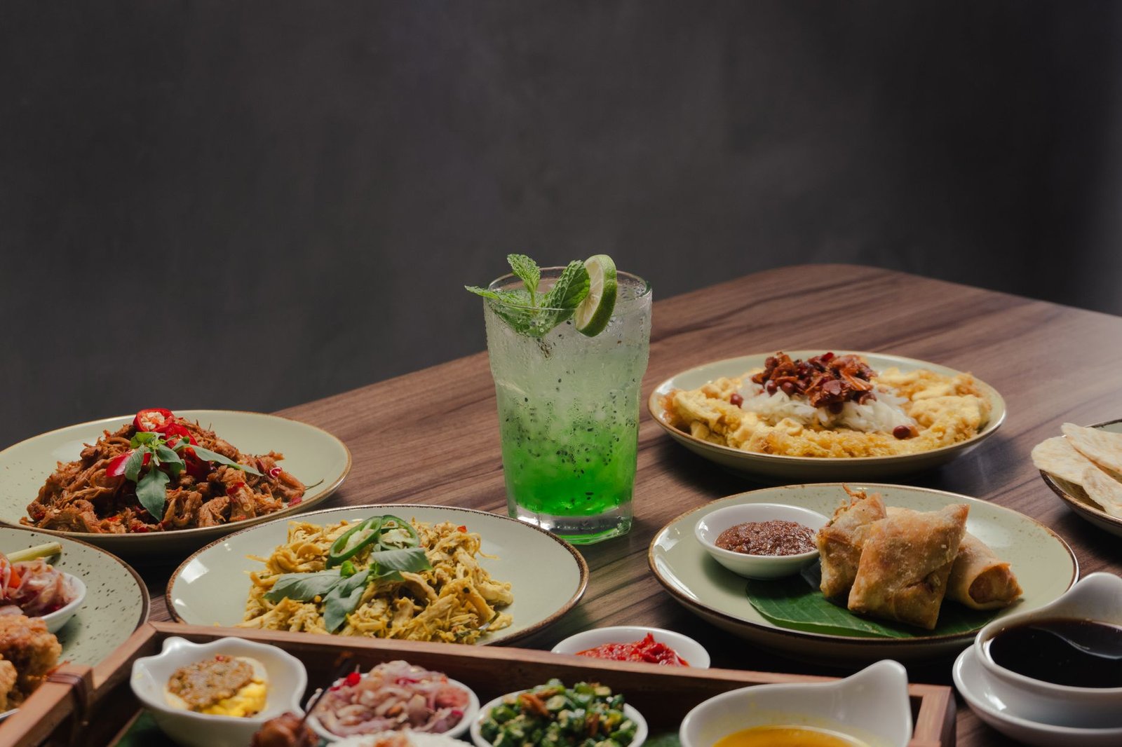 A tempting spread of Asian dishes paired with a refreshing green cocktail on a wooden table.