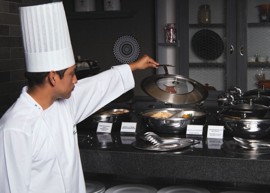 Chef in white uniform opening lid of buffet dish in modern kitchen setting.