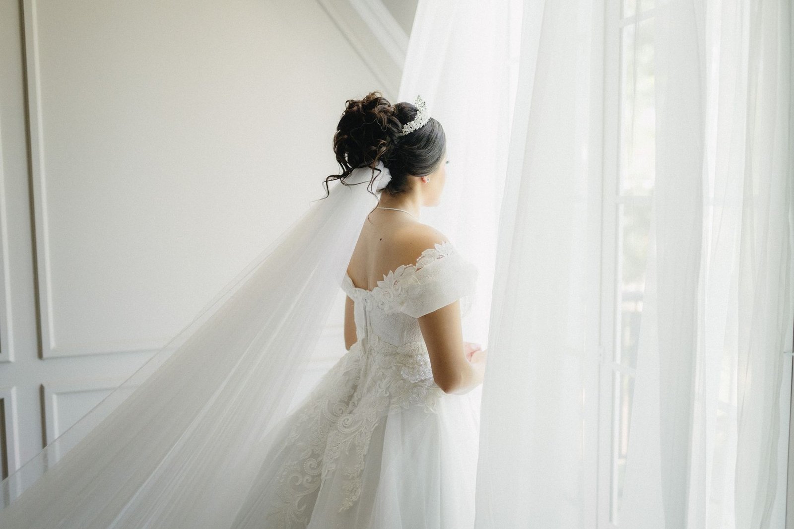 Seorang pengantin wanita yang cantik berpakaian gaun putih dengan penutup kepala melihat keluar dari jendela terang, menangkap momen antisipasi.