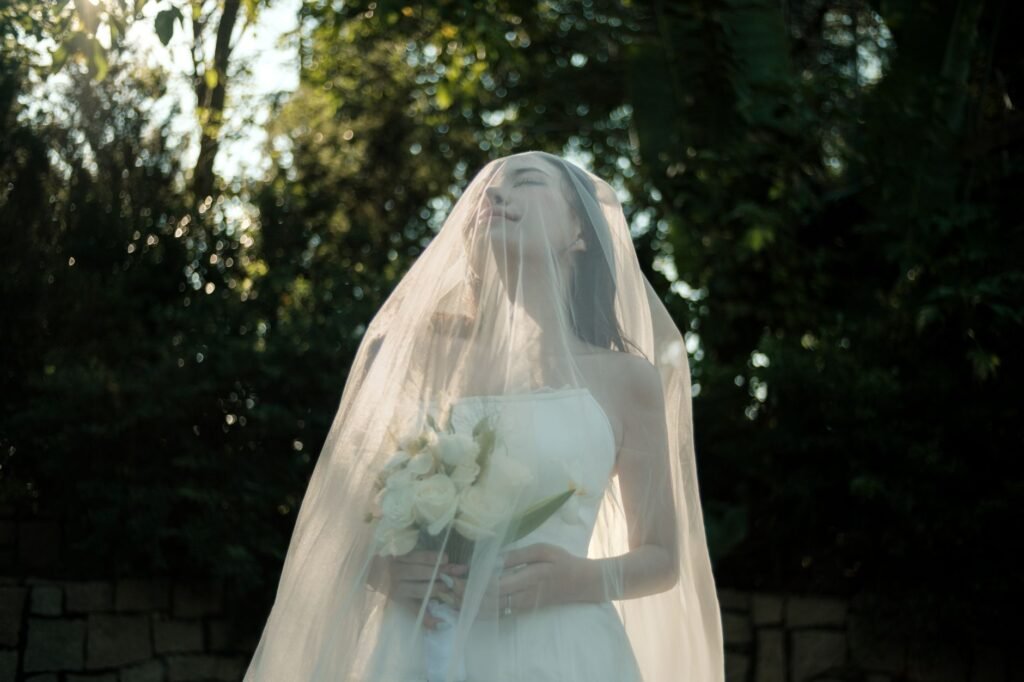 Seorang pengantin wanita yang tenang di hutan yang diterangi sinar matahari, memegang bunga putih, di bawah kerudung yang melambai.