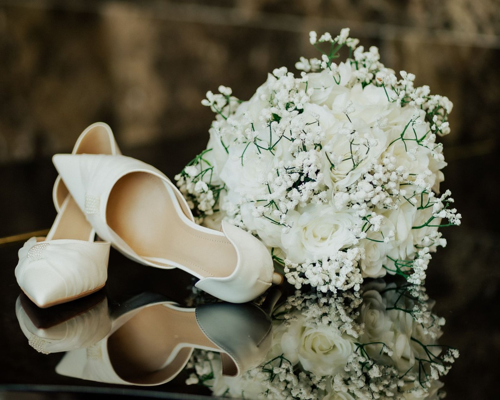 A close-up of elegant white bridal shoes next to a floral bouquet reflected on a shiny surface.