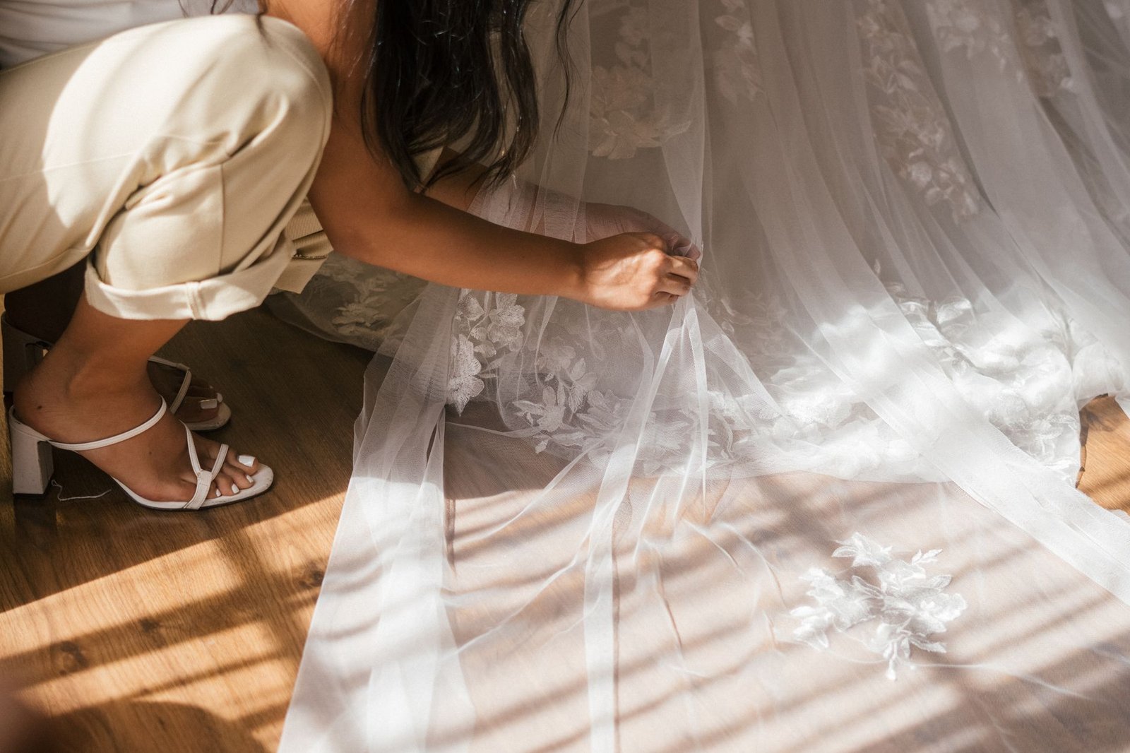 A beautiful bride's veil and dress detail being adjusted indoors, capturing the essence of a wedding day.